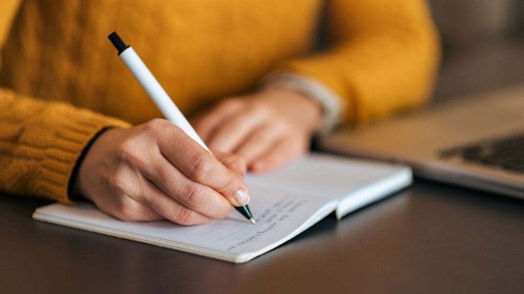 Mão escrevendo metas financeiras em caderno, simbolizando planejamento e organização.
